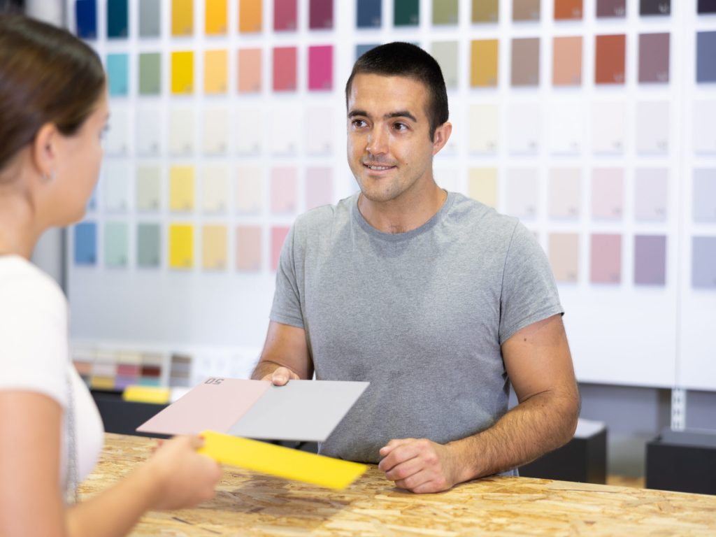 Portrait of positive polite salesman of painting supply store consulting young female client, helping to choose colors of paints for house renovation
